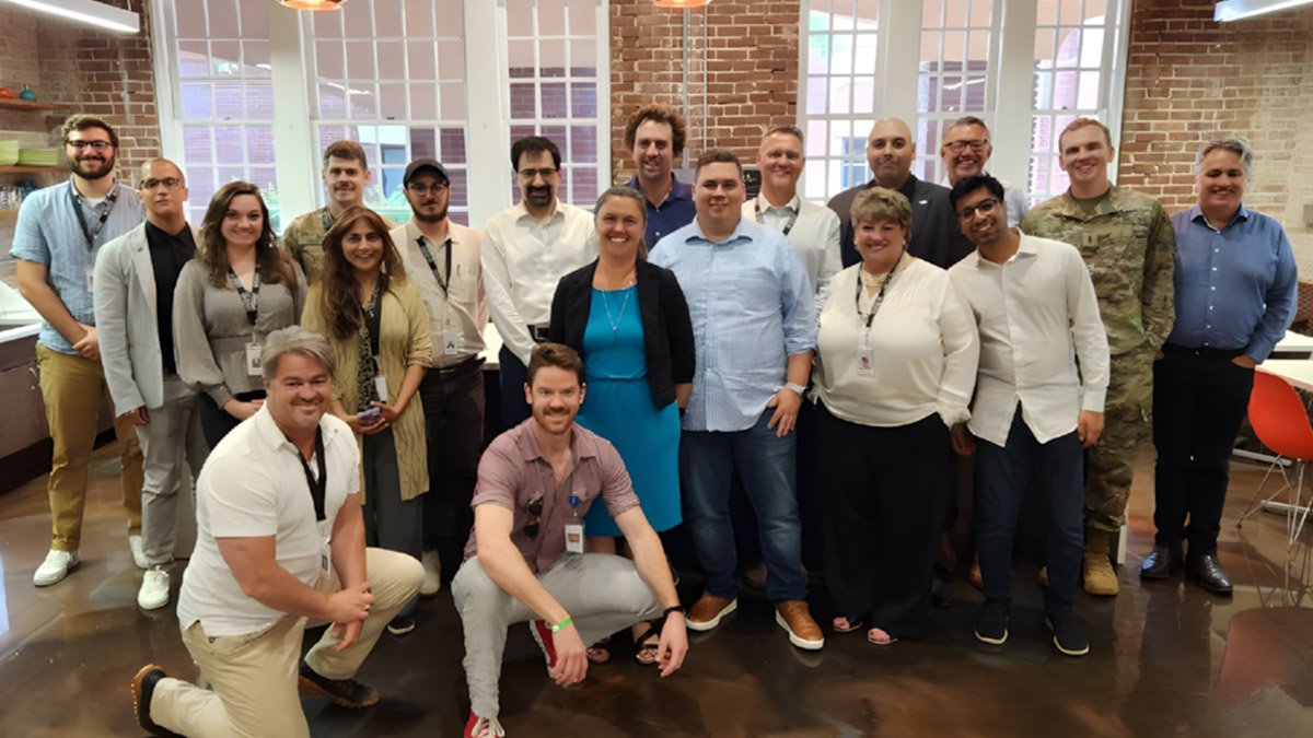 A group of twenty people, including men and women of diverse backgrounds and attire, pose for a photo in a modern, brick-walled kitchen area with large windows and hanging copper lights.