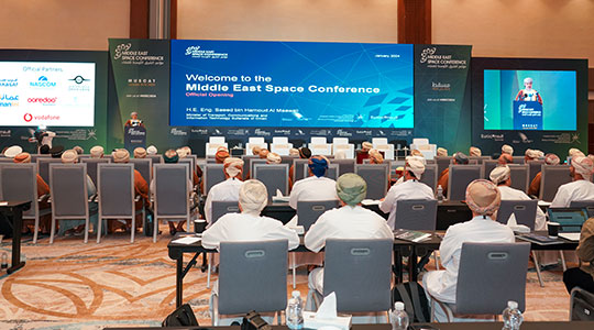 A conference hall with attendees seated and facing a large screen that displays a welcome message for the Middle East Space Conference in Muscat. The stage is adorned with banners featuring the logos of various sponsors and partners.