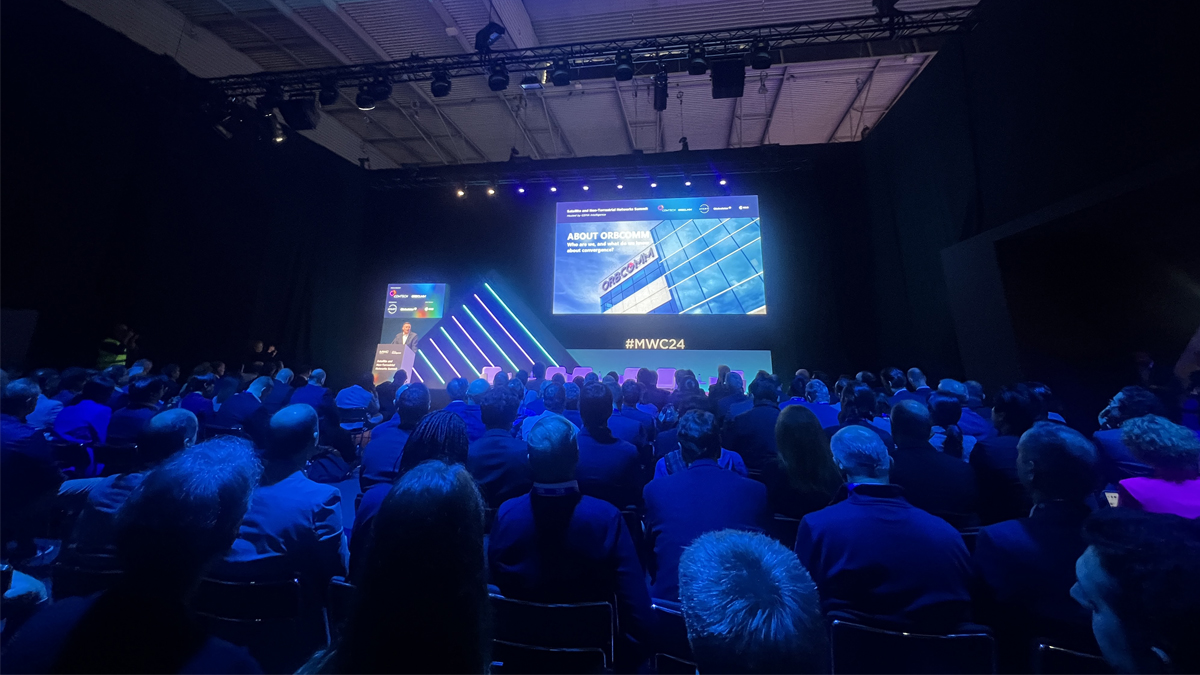 A presenter is speaking at a conference with a slide titled "ABOUT ORBOCOMM" displayed behind, at the Satellite and Non-Terrestrial Networks Summit, with a large audience in attendance.