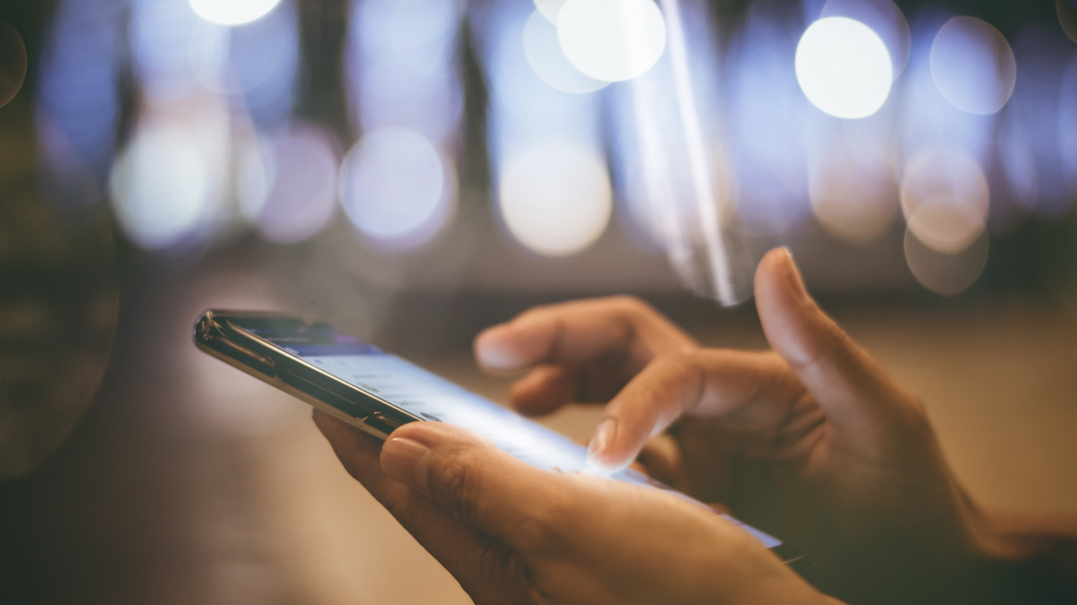 A person's hands holding and interacting with a smartphone with a blurred background of lights.