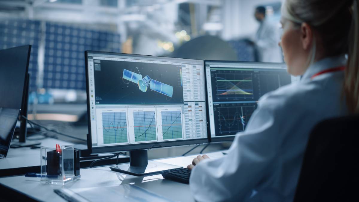 A female engineer analyzes satellite data and calculates orbital trajectories on her computer screens.