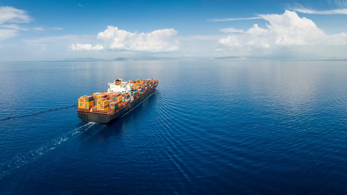 A panoramic aerial view of an industrial cargo container ship traveling across a calm, open sea with distant landmasses and a partly cloudy sky in the background.