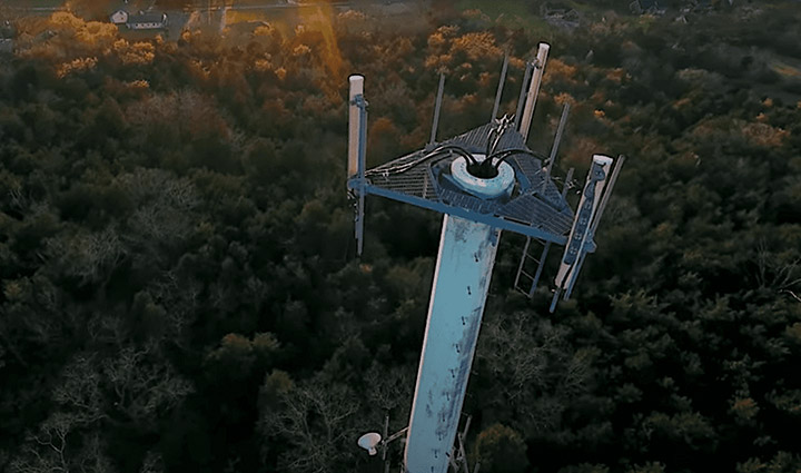 An aerial view of a tall, blue communications tower equipped with antennas and dishes, surrounded by a dense forest at dusk.