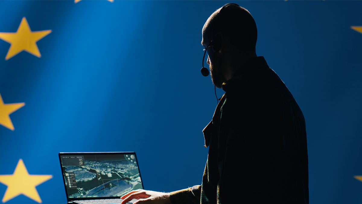 Silhouetted man wearing a headset working on a laptop with a satellite image on the screen, set against a European Union flag background.