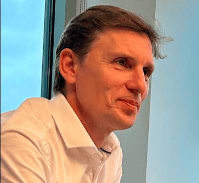 A man in a white shirt is looking to the side, smiling, with a window in the background.