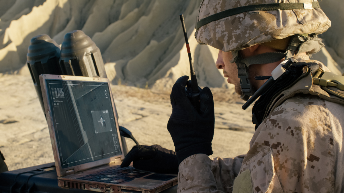 Soldier in camouflage using a rugged laptop and handheld radio in rocky desert terrain, viewing a targeting interface with crosshairs on a vehicle.