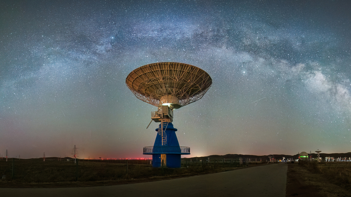 A large radio telescope dish and nearby satellite dishes at a remote observatory beneath a star-filled night sky with the Milky Way visible.