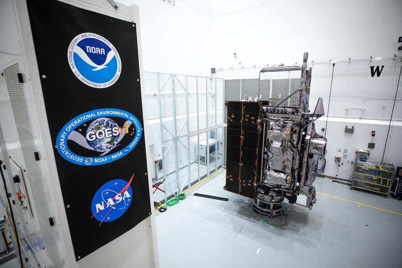 GOES-T satellite inside a cleanroom, wrapped in thermal insulation with solar panels extended and a nearby panel showing NOAA and NASA logos.