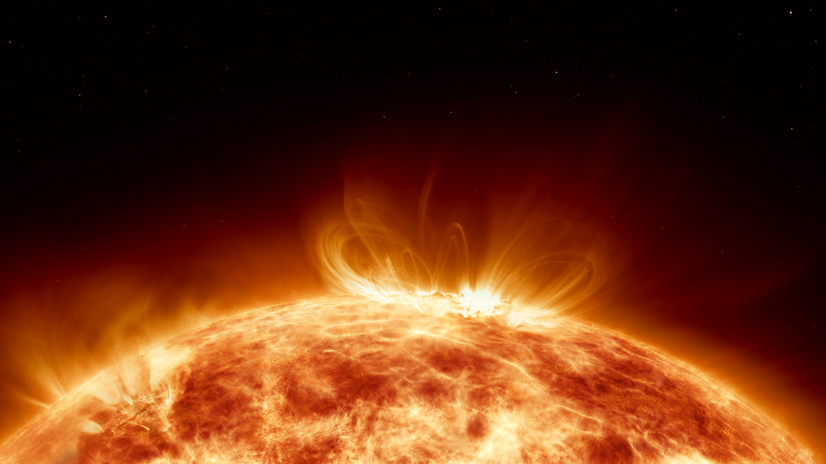 Close-up of the Sun's turbulent, fiery surface with bright coronal loops and prominences arching into space against a star-studded background.