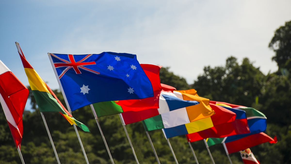 A line of international flags, including the Australian flag, flying outdoors against a sky and trees.