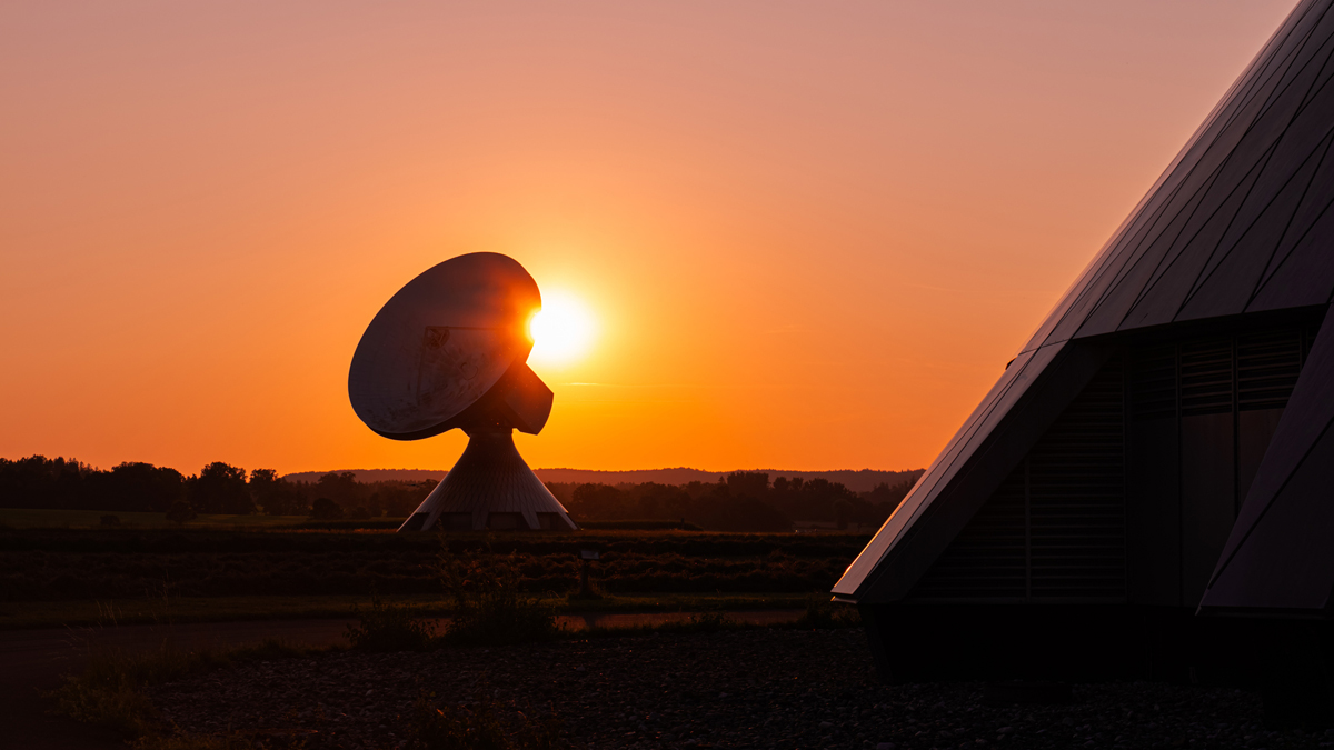 A large satellite ground station dish silhouetted against a vibrant orange sunset, positioned beside a modern, angular building structure.