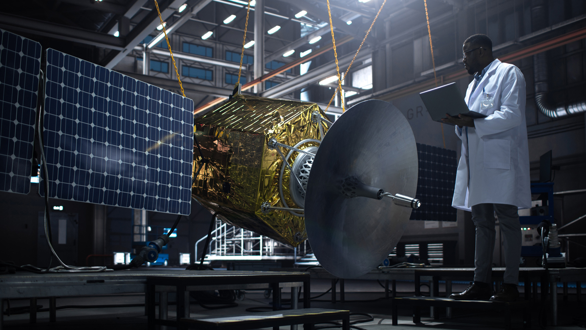 Engineer in a white lab coat holds a laptop while inspecting a gold-bodied satellite with large solar panels and a parabolic antenna suspended by chains inside a high-tech industrial facility.