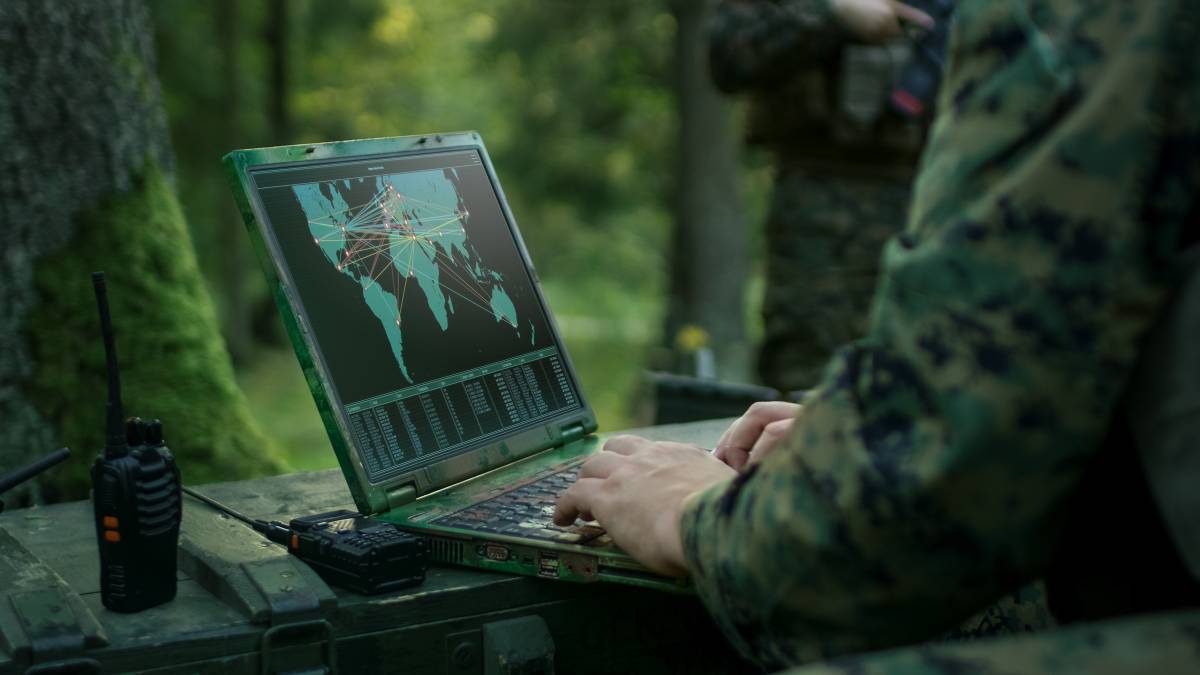 Person in camouflage uniform using a rugged laptop displaying a world map with connections, potentially monitoring global activity, in an outdoor setting.