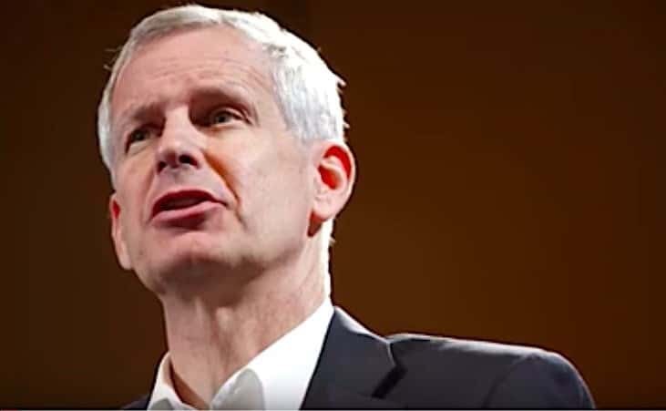 Charlie Ergen, dressed in a dark suit and white shirt, speaks against a dark backdrop.