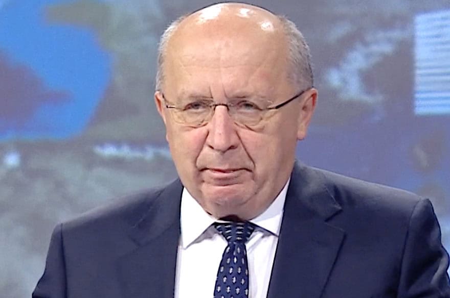 Portrait of Andrius Kubilius in a dark suit, white shirt and patterned tie, wearing glasses and standing before a blurred studio or presentation backdrop.
