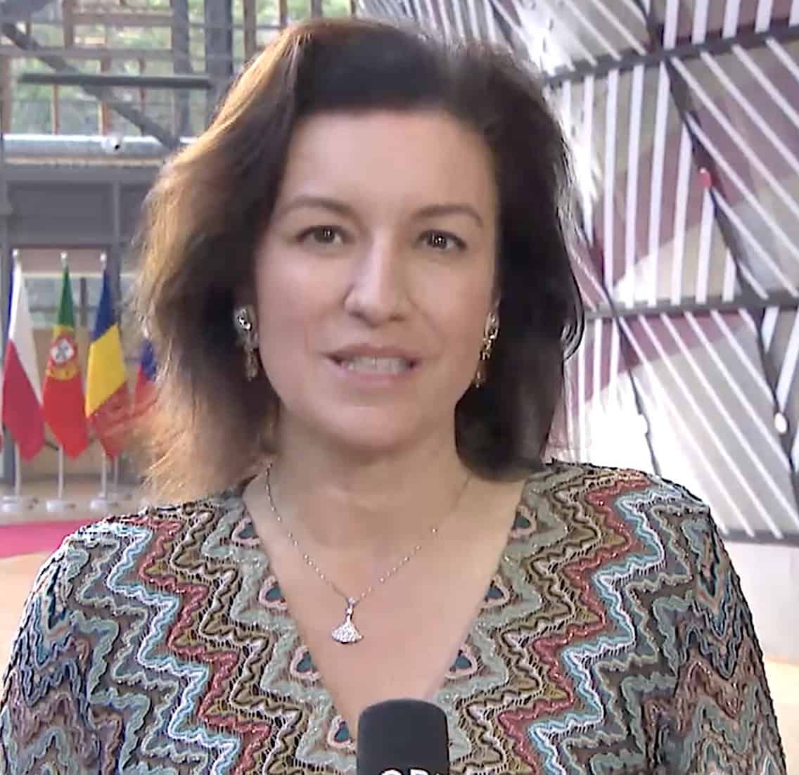 Dorothee Bär speaking into a microphone in a patterned dress, standing before a row of European national flags and a modern glass-and-metal backdrop.