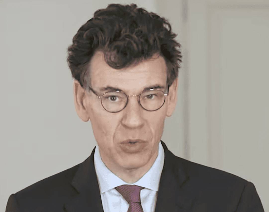 Philippe Baptiste in a dark suit, light blue shirt and maroon tie, wearing round glasses and speaking against a plain light backdrop.