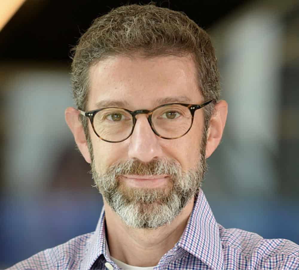 A man with a gray beard and glasses smiles warmly at the camera.