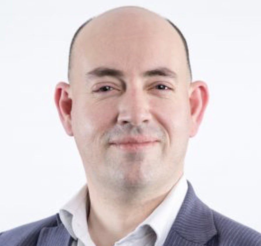 A man with a shaved head smiles at the camera against a white background.