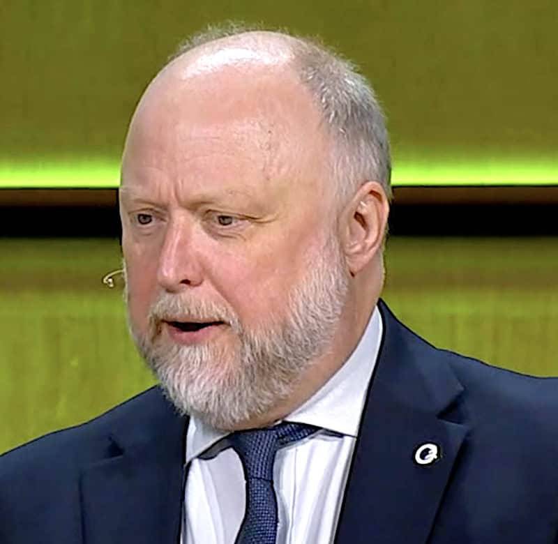 Phil Evans in a dark suit jacket, white shirt and blue tie with a lapel pin, wearing a headset microphone against a greenish-yellow background.