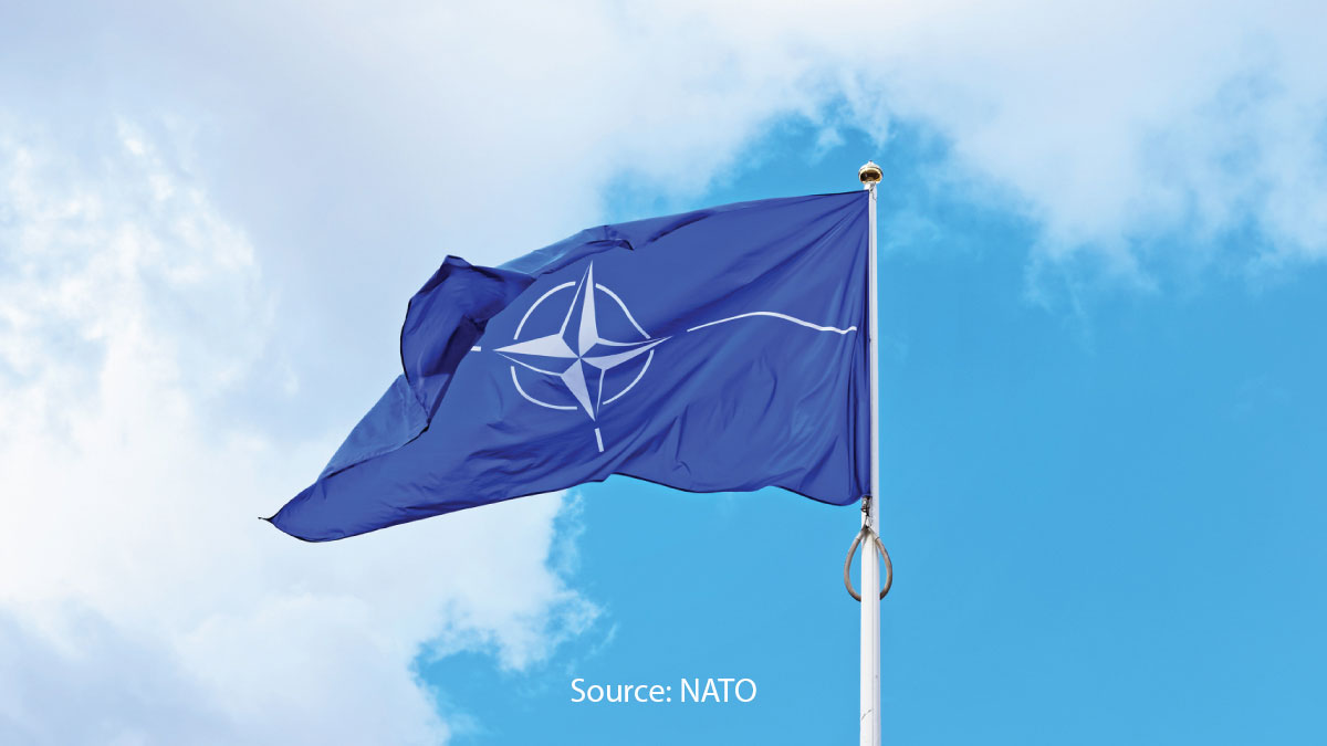 The flag of the North Atlantic Treaty Organization (NATO) waving against a bright, partly cloudy sky.