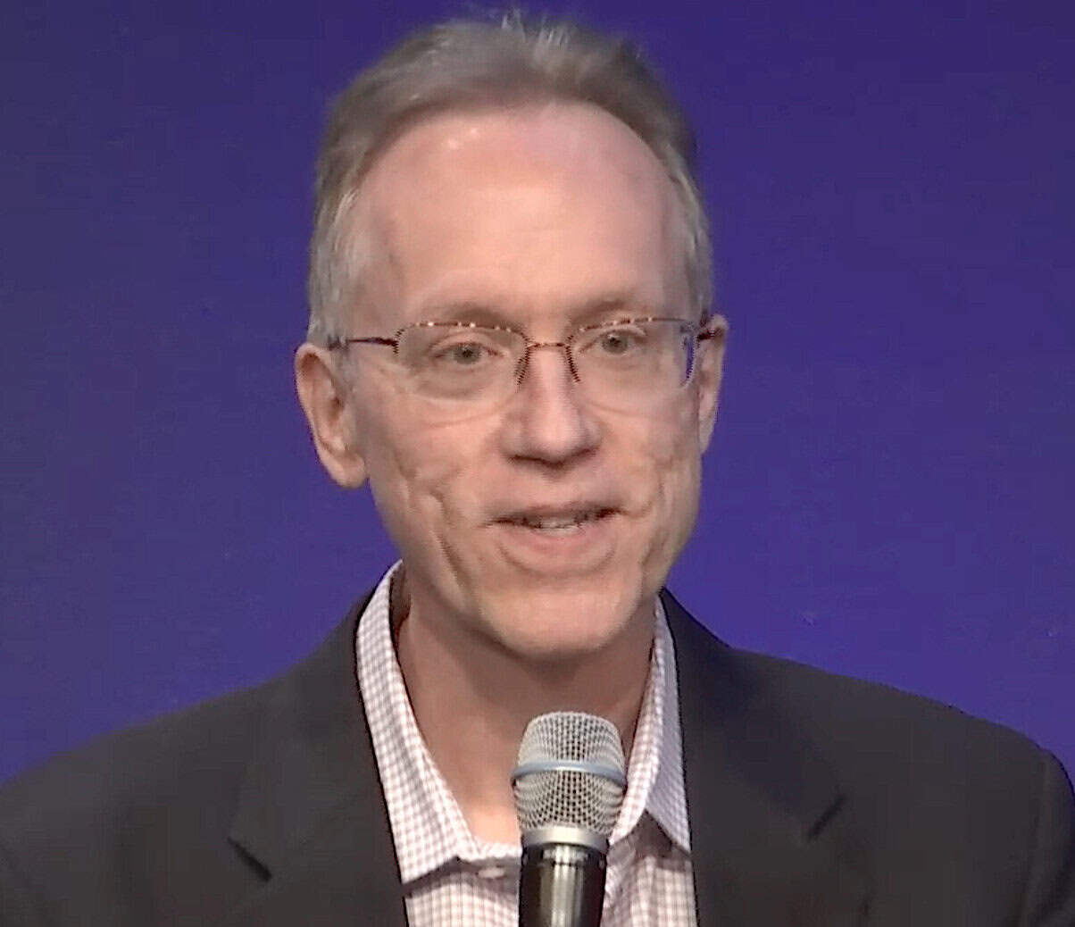 Greg Pelton, Iridium Communications chief technology officer, wearing glasses and a dark blazer over a checkered shirt, speaking into a handheld microphone against a solid blue background.