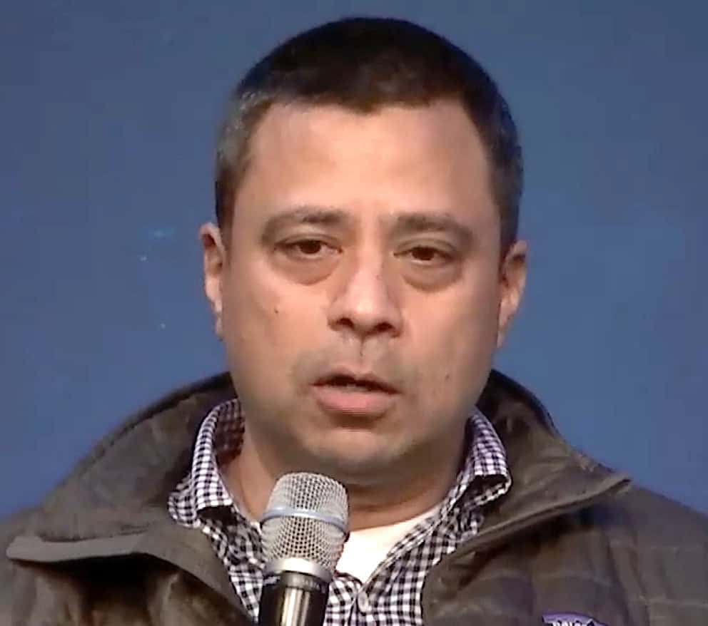 Tarun Gupta, Skylo co-founder and chief product officer, speaking into a handheld microphone while wearing a checkered shirt and dark jacket against a solid blue background.