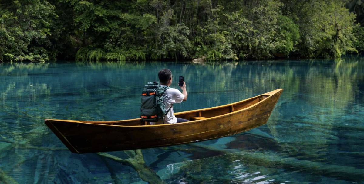 Person in a wooden canoe wearing a white shirt and large green backpack, holding a phone to photograph a crystal-clear lake where submerged logs are visible, surrounded by lush green forest.