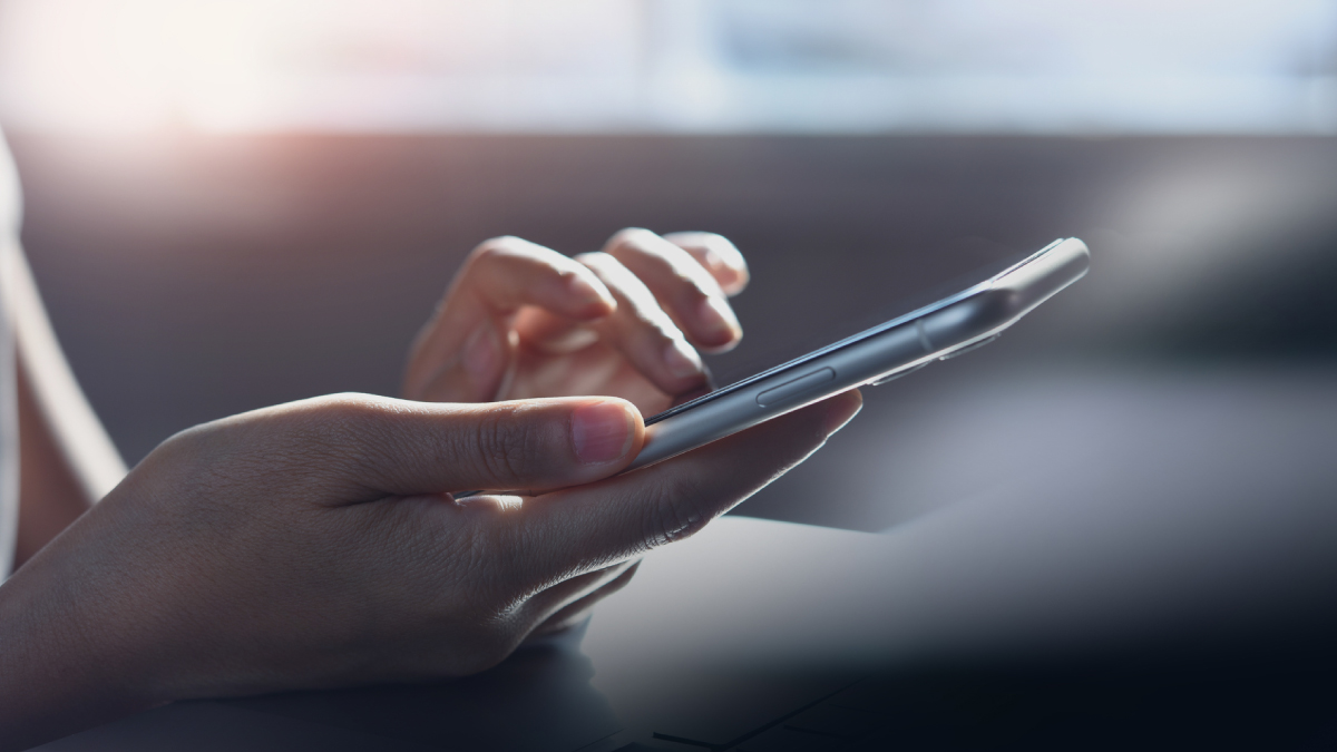 Close-up of a person's hands holding a modern smartphone while the right index finger taps the screen in soft natural window light.