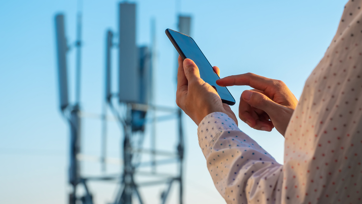 A close-up of a person's hands using a smartphone, with a blurred telecommunications tower in the background against a clear blue sky.