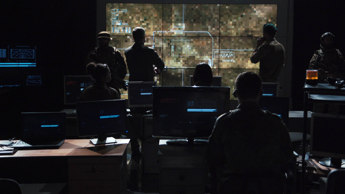 Personnel in a dark command center monitor large digital screens displaying a pixelated map and tactical data.