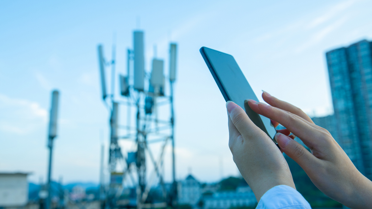 A person holds a smartphone while standing near a cluster of cellular communication antennas against a clear sky.
