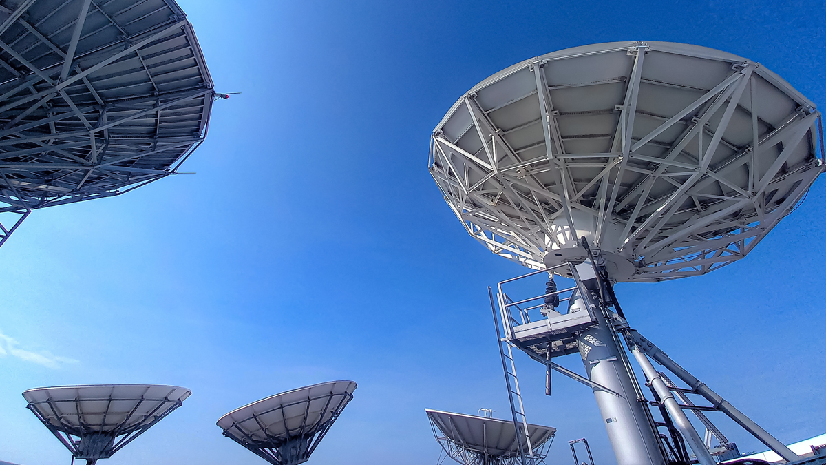 Several large white satellite dishes tilt toward a bright, clear blue sky at a ground station facility.