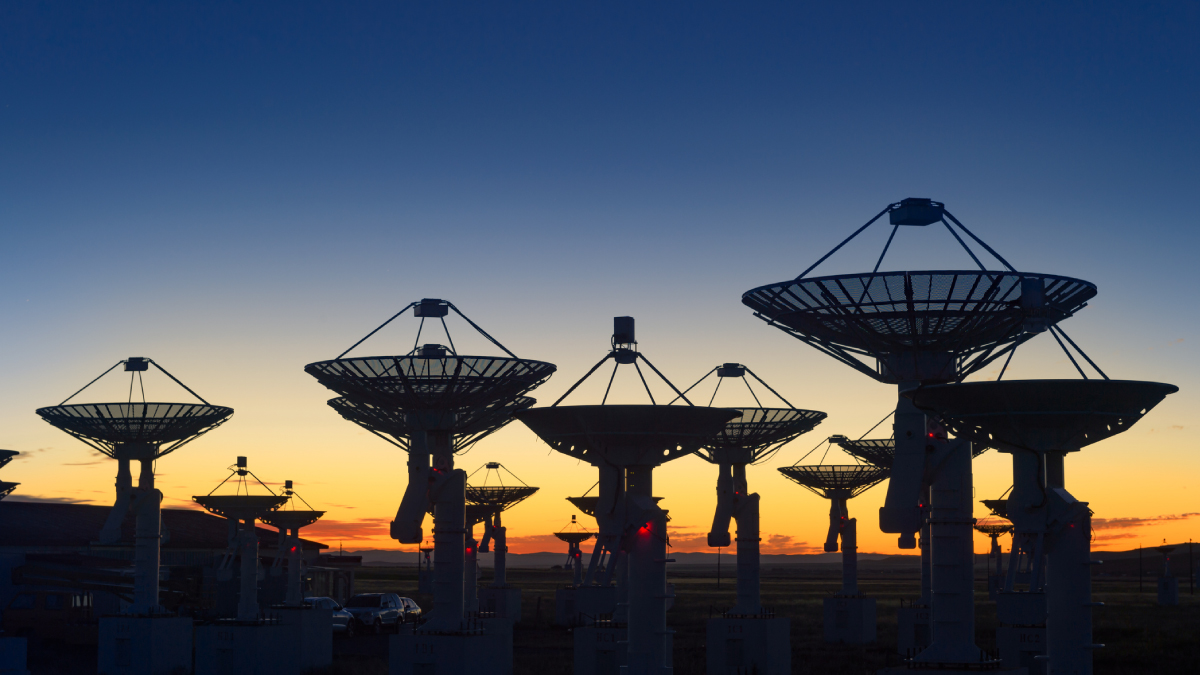 A silhouette of a vast array of satellite dish antennas stretches across a flat landscape under a gradient twilight sky at sunset.