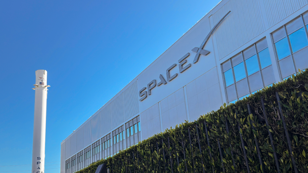 A large white building features the prominent, silver SpaceX logo on its facade under a clear blue sky, with a tall, white rocket booster standing to the left.