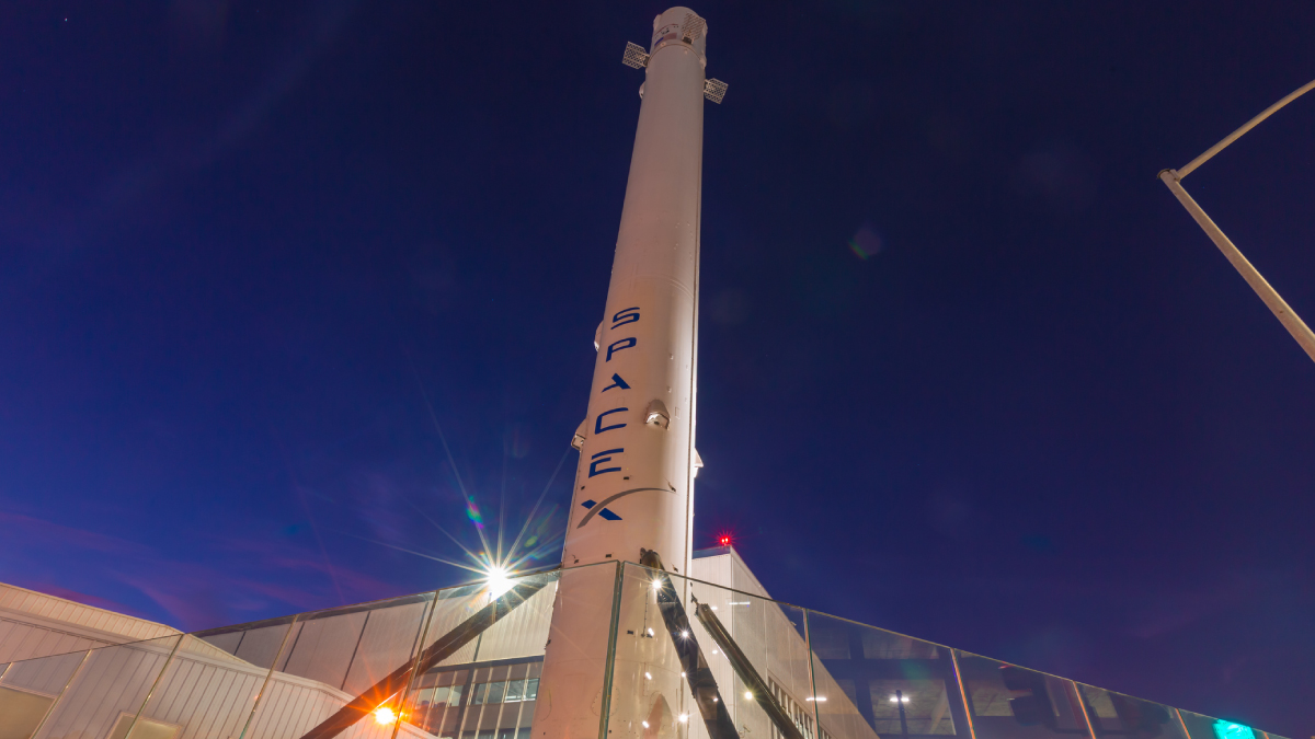 A white SpaceX rocket booster stands vertically against a dark purple and blue night sky, illuminated by bright ground lights.