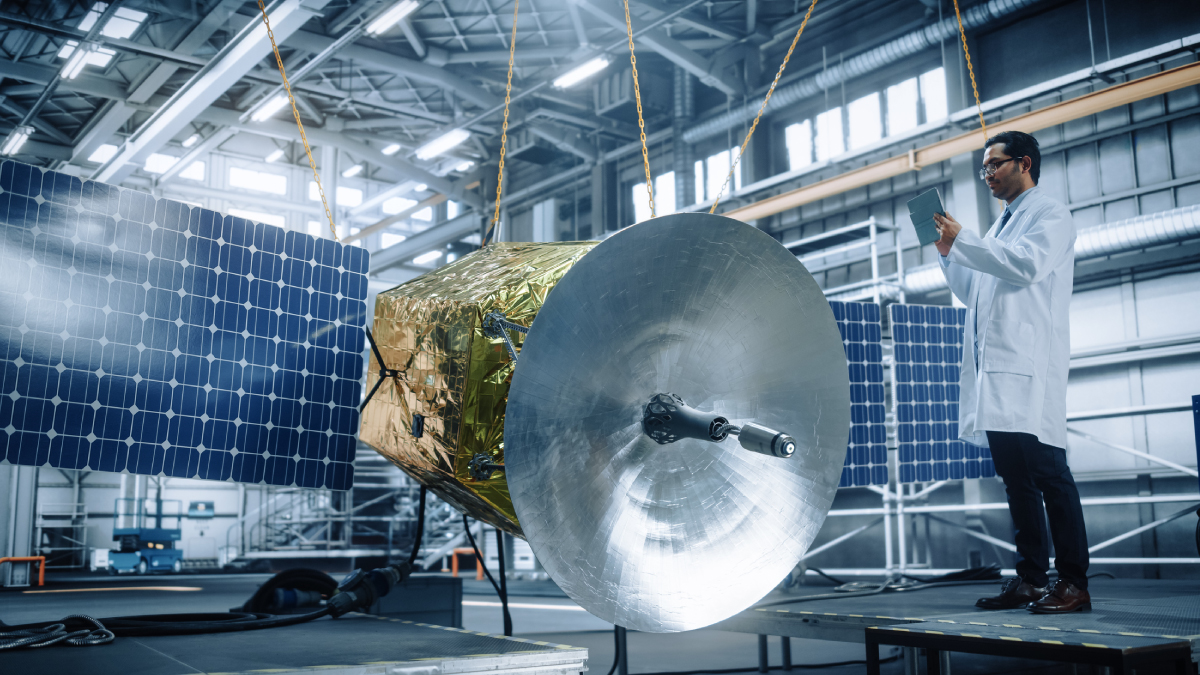 An engineer in a white lab coat uses a tablet to inspect a gold-foil-wrapped satellite with deployed solar panels inside a high-tech manufacturing facility.