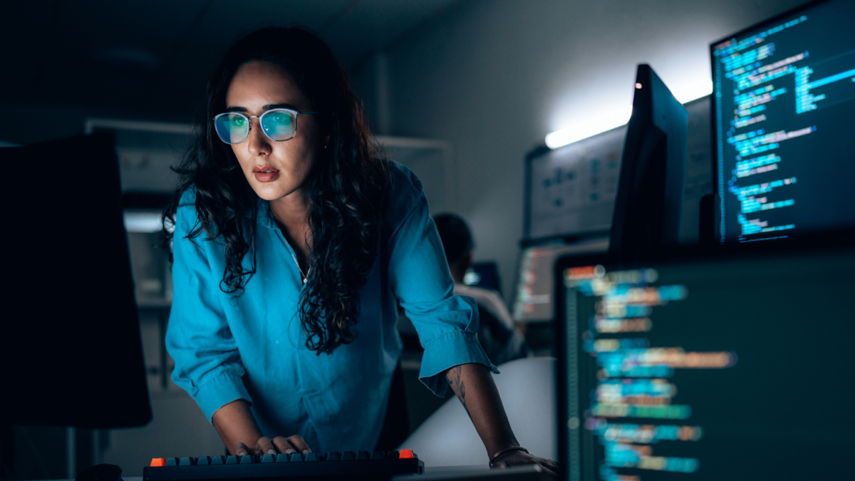 A woman wearing glasses looks intently at multiple computer monitors displaying lines of code in a dimly lit professional setting.
