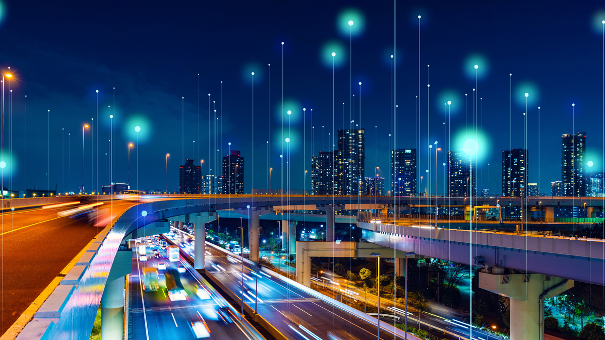 A nighttime cityscape featuring illuminated highway overpasses and moving traffic, overlaid with glowing vertical data lines and nodes to represent smart city telecommunications.