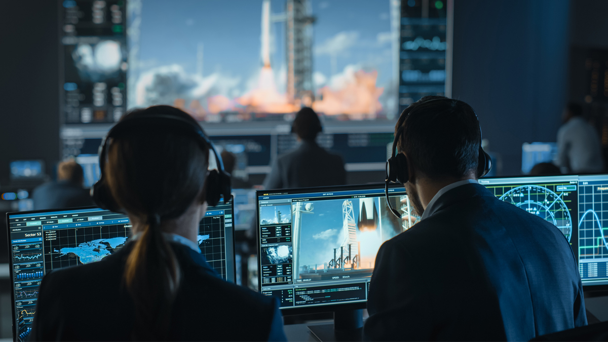 Flight controllers in a mission control center monitor a rocket launch on large screens and desktop monitors.