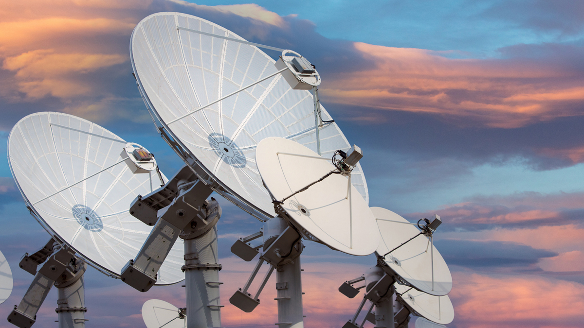 A group of large, white satellite dishes pointing toward a colorful sky with orange and blue clouds at sunset.