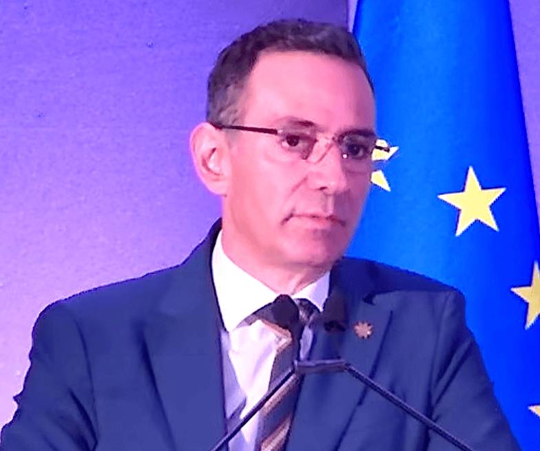 Nicodemos Damianou speaks at a podium with microphones in front of an EU flag and a purple-lit backdrop.