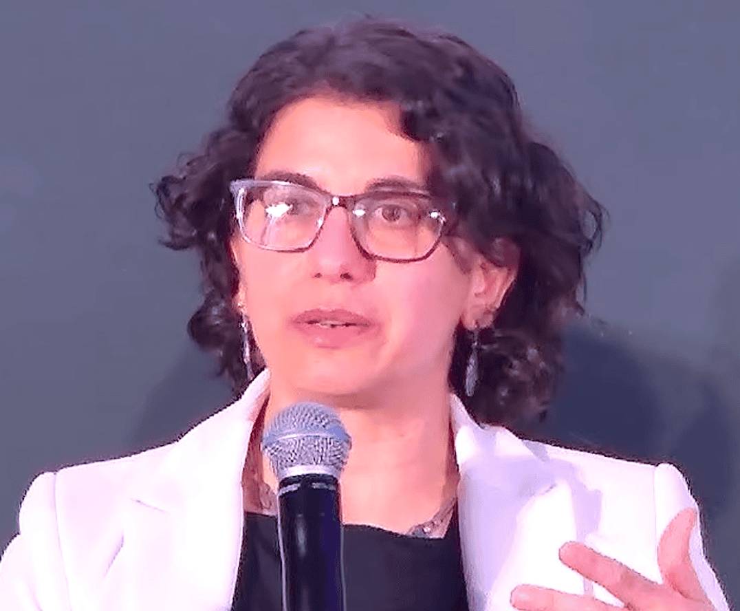 Maria Antonia Ramos Prada in a light-colored blazer is speaking into a handheld microphone against a plain backdrop.