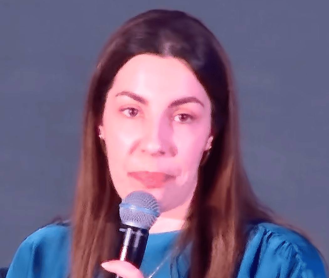 Theodora Liameti holds a microphone while speaking against a plain background.