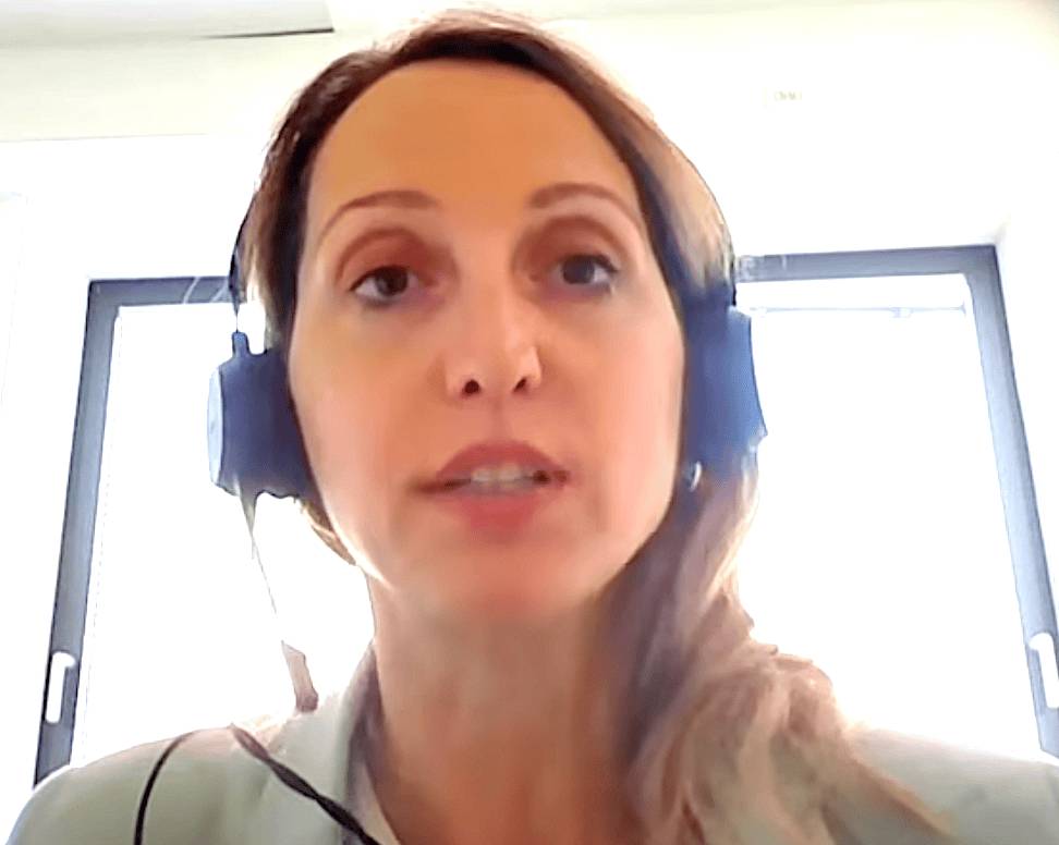 Vicky Kokkori, wearing a headset, is shown indoors with bright light from two windows.
