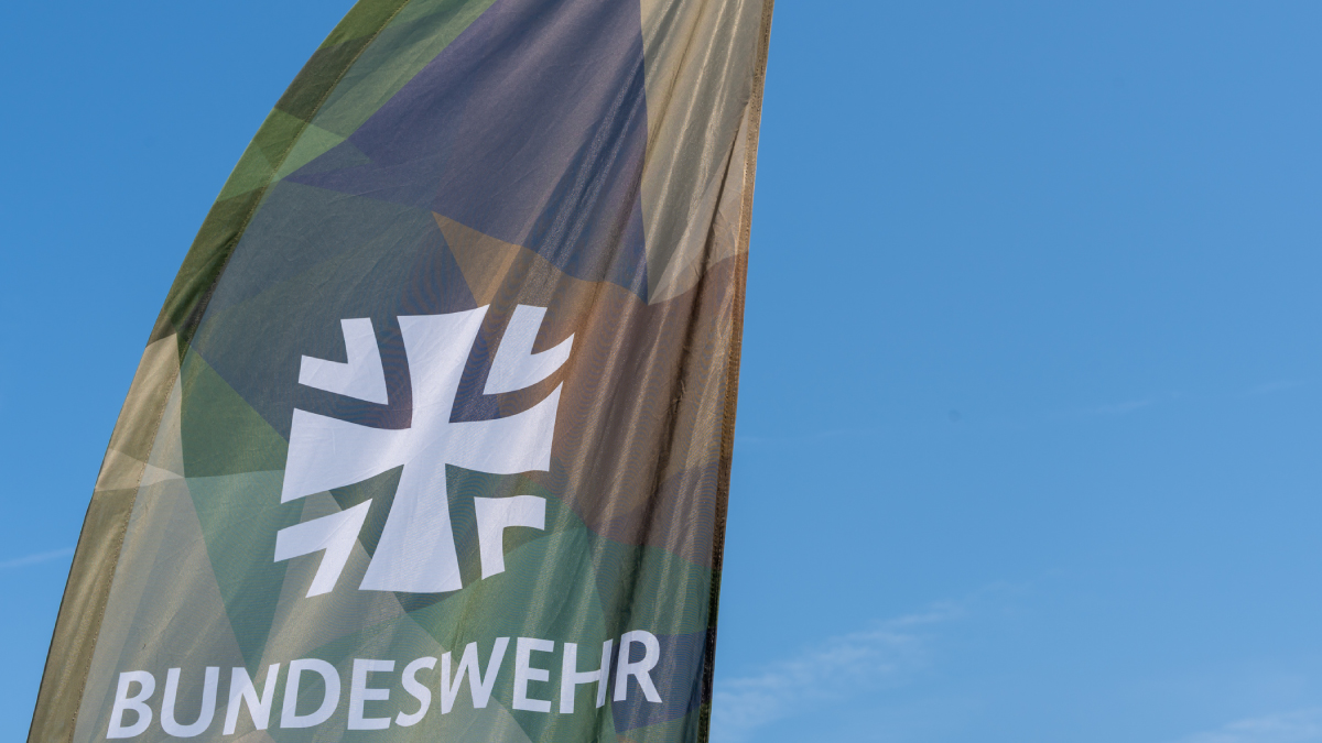 A vertical banner for Germany's armed forces, the Bundeswehr, features their white Iron Cross logo and text over a green and brown geometric camouflage pattern against a clear blue sky.