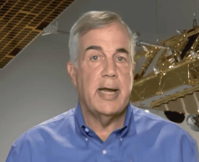 A close-up shot of Iridium CEO Matt Desch wearing a blue button-down shirt, speaking in front of a spacecraft background.