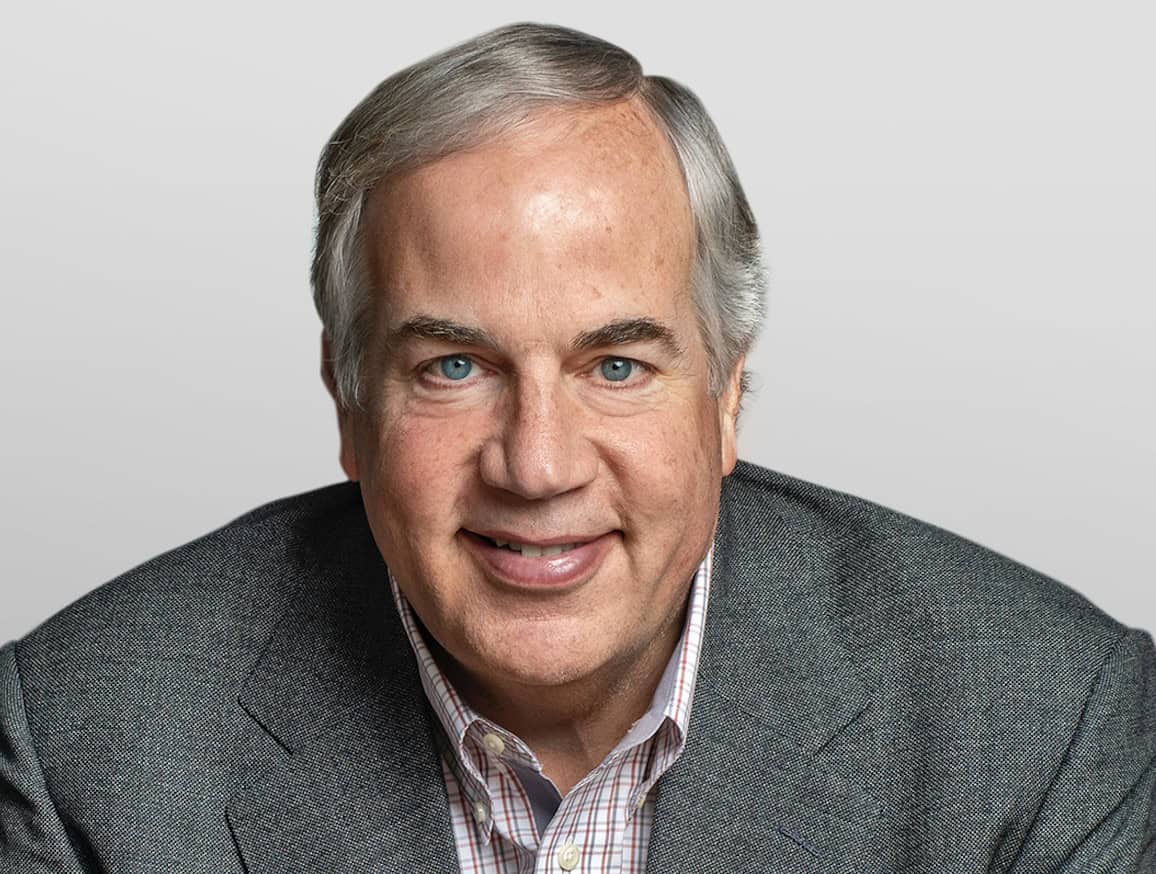 A professional headshot of Matt Desch smiling, dressed in a gray suit jacket and a plaid button-down shirt against a light gray background.
