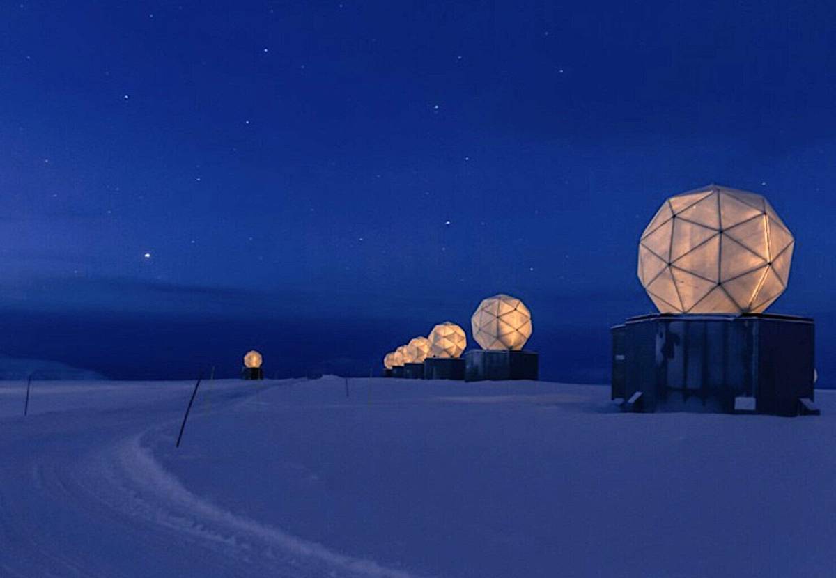 A row of illuminated geodesic ground station domes sits on a snow-covered landscape beneath a dark, starry sky.