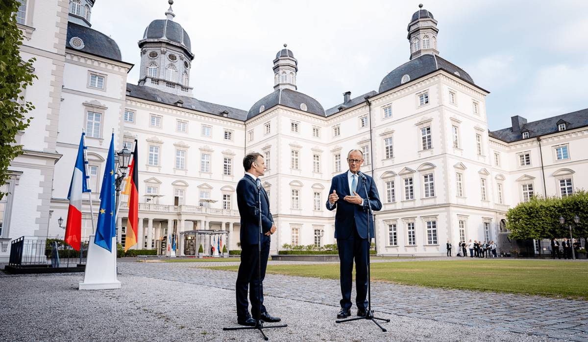 French President Emmanuel Macron and German Chancellor Friedrich Merz stand outdoors at microphones in front of a white, historic palace with the French, European Union, and German flags on display.